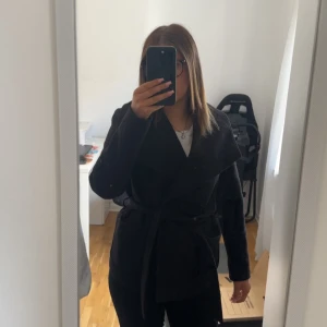 Kappa från vero Moda  - Säljer en svart kappa med bred krage och knytskärp i midjan. Jackan har en stilren look med långa ärmar och stora fickor framtill. Perfekt för dig som gillar en klassisk men trendig stil. Passar till både jeans och kostymbyxor.