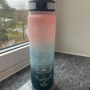 En färgglad vattenflaska  - Bilderna visar en färgglad vattenflaska med motiverande tidsmarkeringar och en praktisk flip-lock-kork. Flaskan har en snygg ombre-design i rosa och blått och ser ut att vara i mycket gott skick utan synliga defekter eller slitage. Perfekt för att hålla koll på vätskeintaget under dagen.