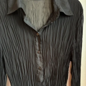 Svart plisserad skjorta från Morgan - Svart långärmad skjorta från Morgan med plisserat tyg och klassisk krage. Skjortan är plisserad, har knappar framtill och är tillverkad i ett tunt, lätt transparent material. Coola manschett-ärmslut.