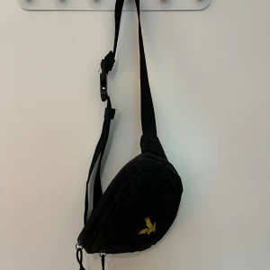 Svart axelväska från Lyle & Scott - Svart axelväska från Lyle & Scott med justerbart band och dragkedja. Väskan har en gul broderad örn på framsidan och är tillverkad i lätt syntetmaterial. Perfekt storlek för mobil, plånbok och nycklar.