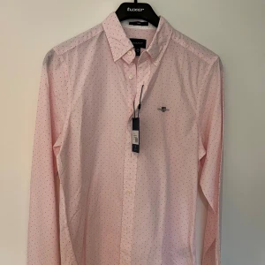 Rosa prickig skjorta från Gant S - Snygg rosa skjorta från Gant i modellen Slim Banker Dot BD. Skjortan har små svarta prickar, broderad logga på bröstet, klassisk krage och knäppning framtill. Tillverkad i bomull och har lång ärm. Perfekt för dig som gillar stilrena detaljer.