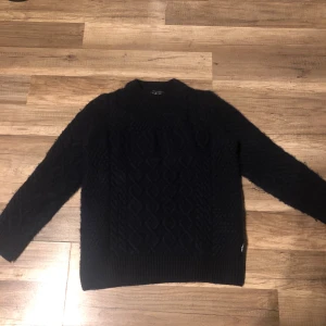 Svart stickad tröja från Barbour XL - Svart stickad tröja från Barbour i storlek XL. Tröjan har ett snyggt kabelstickat mönster och ribbade muddar vid ärmslut och nederkant. Perfekt för dig som gillar klassisk stil med lite edge. Rund hals och långärmad modell.