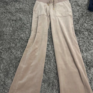Beiga velourbyxor Juicy Couture XS - Supermjuka beiga velourbyxor från Juicy Couture med bootcut passform. Byxorna har bred resår i midjan med snörning, framfickor och bakfickor med guldknappar. Perfekta för en chill och trendig look.