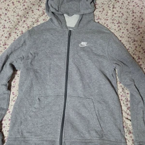 Nike grå kofta  - Grå Nike-hoodie i storlek XL med vit broderad logga på bröstet. Tröjan har huva, hel dragkedja och två stora fickor framtill. Tillverkad i mjuk bomullsmix, perfekt för chill eller träning. Klassisk och enkel design som alltid funkar.
