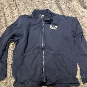 Blå bomberjacka EA7 Emporio Armani - Snygg mörkblå bomberjacka från EA7 Emporio Armani med vit logga på bröstet och coolt vitt band med logga på ryggen. Jackan har hel dragkedja framtill, ribbade muddar och klassisk krage. Perfekt för dig som gillar sportig och stilren look.