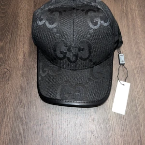 Svart keps med GG-mönster - Snygg svart keps med ikoniskt GG-mönster ton-i-ton över hela kepsen. Kepsen har böjd skärm med läderdetalj och justerbar rem bak i svart läder med guldfärgade detaljer. Perfekt accessoar för en trendig streetstyle-look.