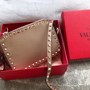 Beige axelväska Valentino med nitar - Säljer en snygg beige axelväska från Valentino Garavani med ikoniska guldfärgade nitar runt kanterna. Väskan är i slät skinn och har justerbar axelrem med nitar. Perfekt för dig som vill ha en stilren men edgy accessoar.(DETTA E BESTÄLLNINGVARA TAR 1-3 VECKOR ATT FÅ HEM DIN PRODUKT) box dustbag får du med!