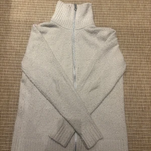 Beige stickad kofta från Brand Island - Mysig beige stickad kofta från Brand Island med hög krage och hel dragkedja framtill. Tröjan har ribbade muddar och en enkel, clean look som passar perfekt till lager-på-lager. Perfekt för kyliga dagar.