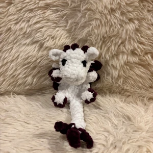 Virkad mini dragon snuggler - Bilderna visar en handgjord, virkad drakleksak i vitt och mörklila garn. Den har svarta ögon och detaljerade ben, svans och vingar. Mjuk och fluffig, perfekt som present eller dekoration. Noggrant och väl utfört arbete utan slitage eller defekter.
