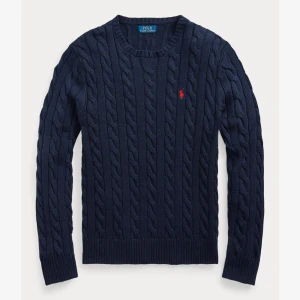 Ralph Lauren cableknitt - Kabelstickad marinblå tröja från Polo Ralph Lauren med rund halsringning och klassisk röd broderad logga på bröstet. Tröjan har ribbade muddar och är tillverkad i mjuk bomull, perfekt för lager-på-lager under kyliga dagar.