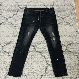 Svarta  jeans med dragkedja - Svarta jeans från Dsquared2 med  passform, slitningar och vita färgstänk. Snygg silverfärgad dragkedja framtill med rund ringdetalj. Jeansen har klassiska fem fickor och en röd Dsquared2-tag vid gylfen.