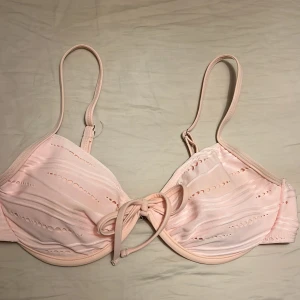 Ljusrosa bikiniöverdel med knyt - Supersöt oanvänd ljusrosa bikiniöverdel med dekorativa hål och knyt framtill. Justerbara axelband och bygel för extra stöd. Tillverkad i mjukt syntetmaterial som torkar snabbt, perfekt för strandhäng eller poolparty.