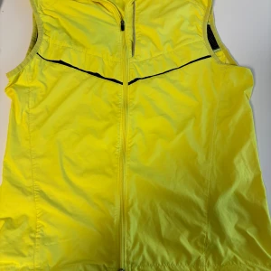 Gul träningsväst från Nike, storlek S - Sportig gul väst från Nike med hel dragkedja framtill och justerbar nederkant med snörning. Västen har svarta detaljer och meshpaneler vid armhålorna för extra ventilation. Perfekt för löpning eller utomhusträning när du vill synas.