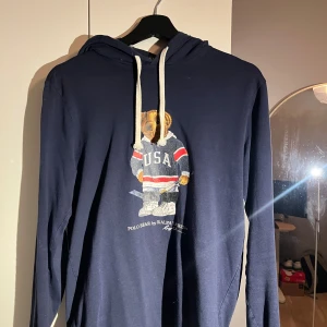 Mörkblå Polo Bear hoodie Ralph Lauren - Säljer en mörkblå hoodie från Ralph Lauren med Polo Bear-print på bröstet. Tröjan har vit snörning i huvan och långa ärmar. Materialet är mjuk bomull och passformen är normal. Perfekt för dig som gillar sportig och chill stil.