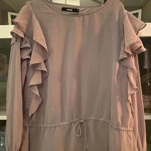Blus - Supersöt ljuslila blus från Bik Bok med volangdetaljer längs ärmarna och knyt i midjan. Blusen har långa ärmar och en lös, flowy passform som ger en romantisk vibe. Perfekt för dig som gillar feminina detaljer och mjuka färger.
