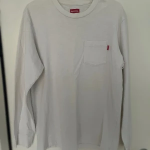 Vit långärmad Supreme tröja - Vit långärmad tröja från Supreme med rund hals och bröstficka med röd Supreme-tag. Tröjan är gjord i mjuk bomull och har en clean, minimalistisk look som passar till streetwear. Perfekt för dig som gillar stilrena plagg med diskret branding.