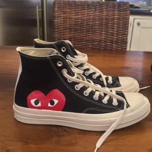 Converse Play Comme des Garçons sneakers - Svarta Converse Chuck Taylor All Star high-top sneakers i samarbete med Comme des Garçons Play. Klassisk vit gummisula, vita snören och tydliga vita sömmar. Ikoniskt rött hjärta med ögon på sidan och Play-logga på innersulan. Tillverkade i bomull. Skorna är helt oanvända.