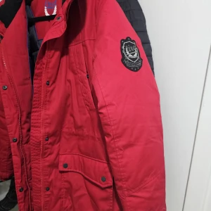 Röd parkajacka från Etirel - Parkajacka i rött från Etirel med svart quiltat foder, huva med fuskpäls och flera fickor med tryckknappar. Jackan har dragkedja och knappar framtill samt en patch på ärmen. Perfekt för kalla dagar och har en schysst streetstil.