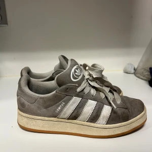 Grå Adidas Campus sneakers - Säljer ett par grå Adidas Campus sneakers med klassiska vita ränder på sidan och logga på plösen. Skorna har snörning, rund tå och platt sula. Ovandelen är i mjuk mocka och insidan är fodrad för extra komfort. Perfekt för dig som gillar retrostil och streetwear. Strl 41 och en halv