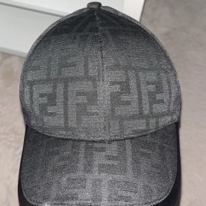 Svart Fendi keps med logomönster - Snygg svart keps från Fendi med det klassiska FF-logomönstret över hela kepsen. Kepsen har böjd skärm och justerbar passform. Tillverkad i ett slitstarkt syntetmaterial som känns lyxigt och trendigt. Perfekt för dig som vill ha en exklusiv streetstyle-look.