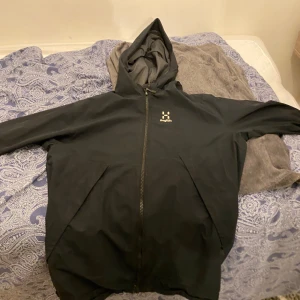 Svart vindjacka från Haglöfs med huva - Svart vindjacka från Haglöfs med praktisk huva och hel dragkedja framtill. Köpt för 2700 kr på Intersport priset kan diskuteras vid snabb affär. Använt i 4 månader.