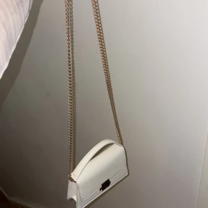 Vit axelväska med kedja - Snygg vit axelväska med stilren design och guldfärgad kedja som axelrem. Väskan har ett elegant metallspänne framtill och är tillverkad i slätt skinnmaterial. Perfekt storlek för det viktigaste och passar till många olika outfits. Lite smutsig inuti men inget som stör. 