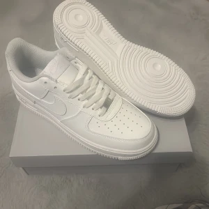 Nike Air Force 1 helvita sneakers - Helt nya och oanvända Nike Air Force 1 sneakers i helvit färg med klassisk låg siluett. Skorna har rund tå, snörning och perforerad tåbox för extra ventilation. Tillverkade i skinn med vit yttersula och ikonisk Nike Swoosh på sidan. Perfekt för dig som gillar clean och tidlös streetstil.