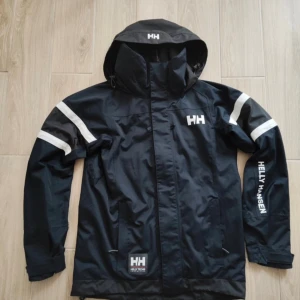 Svart vindjacka/regnjacka från Helly Hansen - Snygg svart vindjacka från Helly Hansen med vita detaljer och logga på bröstet och ärmen. Jackan har huva, dragkedja och två fickor framtill. Materialet är slitstark polyester och jackan fyller en hel del funktioner, kan användas som skidjacka, regnjacka, vindjacka vid segling osv.