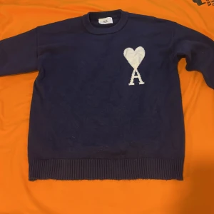 Mörkblå stickad tröja från Ami - Mörkblå stickad tröja från Ami med rund halsringning och ribbade muddar. På bröstet finns ett vitt hjärta och bokstaven A som snygg detalj. Perfekt för dig som gillar stilrena plagg med en twist.