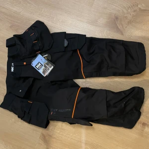 Svarta arbetsbyxor Helly Hansen C48 - Svarta arbetsbyxor från Helly Hansen med orange detaljer och flera praktiska fickor. Tillverkade i slitstarkt Cordura-material, perfekt för dig som vill ha både funktion och stil på jobbet. Snygg logga på benet och coola kontrastdetaljer.
