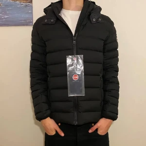 Svart pufferjacka från Colmar - Svart pufferjacka från Colmar med avtagbar huva och diskret logga på ärmen. Jackan har en quiltad design, dragkedja framtill och två sidofickor med dragkedja. Perfekt för kalla dagar och har en clean, modern look. Storlek:S