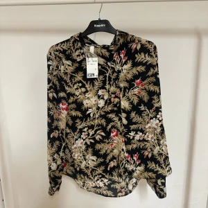 Svart blommig skjorta från H&M - Svart skjorta från H&M med blommigt mönster i beige, vitt och rött. Skjortan har klassisk krage, knappar framtill och lång ärm. Materialet är mjukt och lätt, perfekt för dig som gillar detaljer och färg. Snyggt flow och relaxed fit.