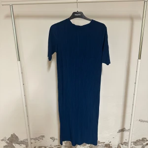 Marinblå långklänning från KappAhl - Säljer en stilren marinblå långklänning från KappAhl med plisserad struktur. Klänningen har rund halsringning och korta ärmar, vilket ger en enkel men modern look. Perfekt för dig som gillar minimalistisk stil och djupa färger.