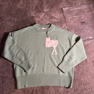 Mintgrön stickad tröja från Ami - Snygg mintgrön stickad tröja från Ami Alexandre Mattiussi med ribbade muddar och nederkant. På bröstet finns ett stort vitt hjärta med bokstaven A. Tröjan har rund hals och lång ärm, perfekt för dig som gillar stilrena detaljer och mjuka material.