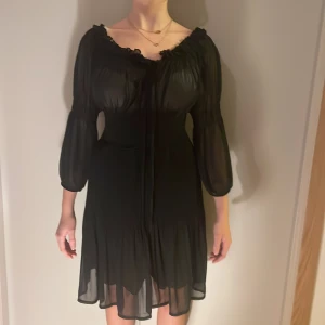 Svart offshoulder klänning Zara - Svart kortklänning från Zara Basic med offshoulder och trekvartsärmar. Klänningen har volangdetaljer vid halsringningen och är tillverkad i ett lätt, transparent polyestertyg med smockad midja för snygg passform. Perfekt för dig som gillar romantisk och trendig stil.