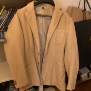 Beige kavaj från Faconnable - Snygg beige kavaj från Faconnable, tillverkad i Italien. Klassisk modell med tre fickor framtill och knappar vid ärmslut. Tidlös design i mjukt material, perfekt för dig som gillar stilrena plagg med exklusiv känsla.