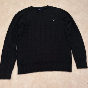 Svart kabelstickad tröja från Gant - Svart kabelstickad tröja från Gant i premium bomull. Klassisk rund halsringning, ribbade muddar och diskret broderad logga på bröstet. Perfekt för dig som gillar stilren och tidlös design. Priset går alltid att diskutera. Hör av er om ni undrar över något 😊