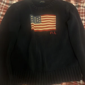 Mörkblå stickad tröja Polo Ralph Lauren - Säljer en mörkblå stickad tröja från Polo Ralph Lauren med amerikansk flagga och RL-broderi på bröstet. Tröjan har rund hals, långa ärmar och breda ribbade muddar. Perfekt för dig som vill ha en klassisk och stilren look.