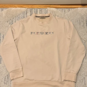 Beige Burberry sweatshirt med logga - Snygg beige sweatshirt från Burberry med klassisk logga framtill i rutigt mönster. Tröjan har rund hals, långa ärmar och ribbade muddar. Perfekt för dig som gillar stilrena och exklusiva plagg med en touch av lyx.