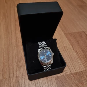 Seiko herrklocka med blå urtavla - Säljer min Saiko mod-klocka som är byggd i stil med en Datejust. Den har en elegant mörkblå/marinblå urtavla och ett klassiskt Jubilee-armband som ger en riktigt tidlös känsla. Klockan är 39 mm i diameter och har självlysande visare samt tim- och minutmarkeringar, vilket gör den lätt att läsa även i mörker. Den har burits i cirka två veckor och är därför i princip som ny, med endast något enstaka litet spår av användning. Seiko NH35A urverk. Klockan levereras med en box och fungerar felfritt.