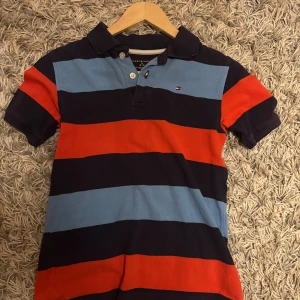 Randig pikétröja Tommy Hilfiger M - Snygg pikétröja från Tommy Hilfiger med breda ränder i marinblått, ljusblått och rött. Klassisk krage, korta ärmar och knappar framtill. Perfekt för en avslappnad och sportig look.