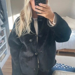 Svart jacka från H&M  - Säljer en svart pälsjacka med cool snörning och knappar framtill. Jackan har en rak och loose passform med långärmade ärmar och mjukt pälsmaterial som ger en lyxig vibe. Perfekt för dig som vill ha en statement piece i garderoben.