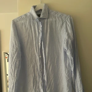 Massimo Dutti Ljusblå randig linneskjorta - Snygg ljusblå skjorta med tunna vita ränder och klassisk krage. Skjortan har långa ärmar och knäppning framtill med vita knappar.  Använd bara ett par gånger