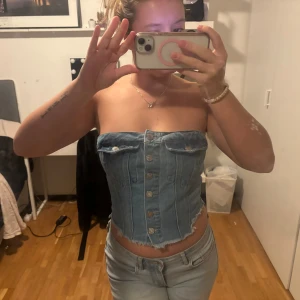 Blå denim tubtopp med knappar - Säljer en snygg blå tubtopp i denim med knappar framtill och fransad nederkant. Toppen har två dekorativa fickor med knapp och smockad rygg för skön passform. Perfekt för dig som vill ha en cool och trendig look.