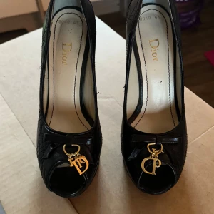 Svarta Dior pumps med gulddetaljer - Säljer ett par svarta pumps från Dior i skinn med hög klack och plattform. Skorna har öppen tå, quiltad yta och snygga guldfärgade Dior-hängen framtill. Röd sula ger extra edge och lyxig känsla. Perfekt för dig som vill sticka ut med exklusiva detaljer. Klackhöjd 13,5 cm