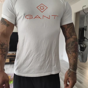 Vit GANT t-shirt med röd logga - Snygg vit t-shirt från GANT med röd logotyp tryckt på bröstet. Klassisk rund hals och korta ärmar. Skön passform och tillverkad i mjuk bomull som känns fräsch mot huden. Perfekt för dig som gillar stilrena och enkla plagg med tydlig branding.