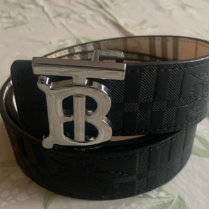 Burberry vändbart bälte med TB-spänne - Snyggt vändbart bälte från Burberry med klassiskt rutigt mönster på ena sidan i beige, rött och svart samt svart skinn med präglat mönster på andra sidan. Stort silvrigt TB-spänne framtill. Perfekt accessoar för att lyfta din outfit. Jag har en till storlek 115cm