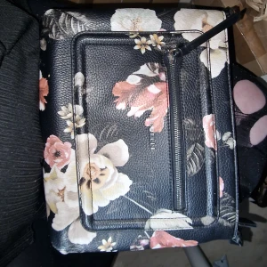 Blommig svart axelväska från Fiorelli - Snygg svart axelväska från Fiorelli med blommigt mönster i beige, rosa och vitt. Väskan har en praktisk dragkedja framtill och är gjord i skinnimitation med struktur. Perfekt storlek för det viktigaste och har en modern vibe.