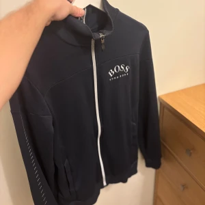  Hugo Boss tröja  - Snygg svart ziptröja från Hugo Boss med vit dragkedja och logga på bröstet. Tröjan har hög krage med Hugo Boss-detalj, långa ärmar och praktiska fickor. Perfekt för en stilren och sportig look.