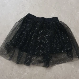 Kjol - Supersöt svart kortkjol från Mywear Baby med flera lager tyll och små guldglittriga prickar över hela kjolen. Resår i midjan för skön passform och ett mjukt innerfoder. Perfekt för dig som vill sticka ut lite extra med glitter och volym.